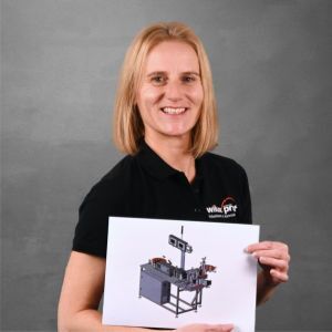 Angela Büchel, CAD Konstrukteurin bei WILUX PRINT, hält einen technischen Entwurf einer Maschine in den Händen, während sie in einem schwarzen Polohemd mit Firmenlogo vor einem neutralen Hintergrund lächelt.