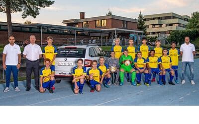 Gruppenfoto der Juniorenmannschaft Cb des FC Hinwil mit ihren Trainern und Sponsoren, unterstützt von WILUX PRINT, aufgenommen vor einem Vereinsgebäude.