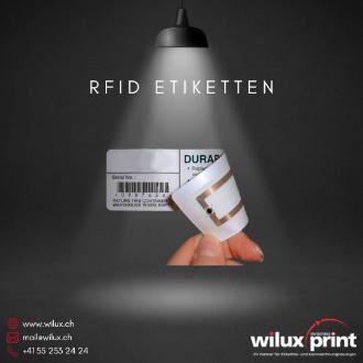 Eine Hand hält ein RFID-Etikett, das einen integrierten Funk-Chip (Transponder) zeigt. Im Hintergrund ist ein weiteres RFID-Etikett mit Barcode sichtbar.