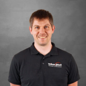Marcel Lattmann, Teil des Teams WILUX PRINT, Leiter Forschung & Entwicklung, Montage, Produktion und Logistik, trägt ein schwarzes Poloshirt mit WILUX PRINT Logo, vor einem neutralen Hintergrund.