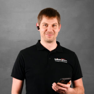 Marcel Lattmann, Teil des Teams WILUX PRINT, Leiter Forschung & Entwicklung, Montage, Produktion und Logistik, mit Headset und Smartphone, trägt ein schwarzes Poloshirt vor einem neutralen Hintergrund.